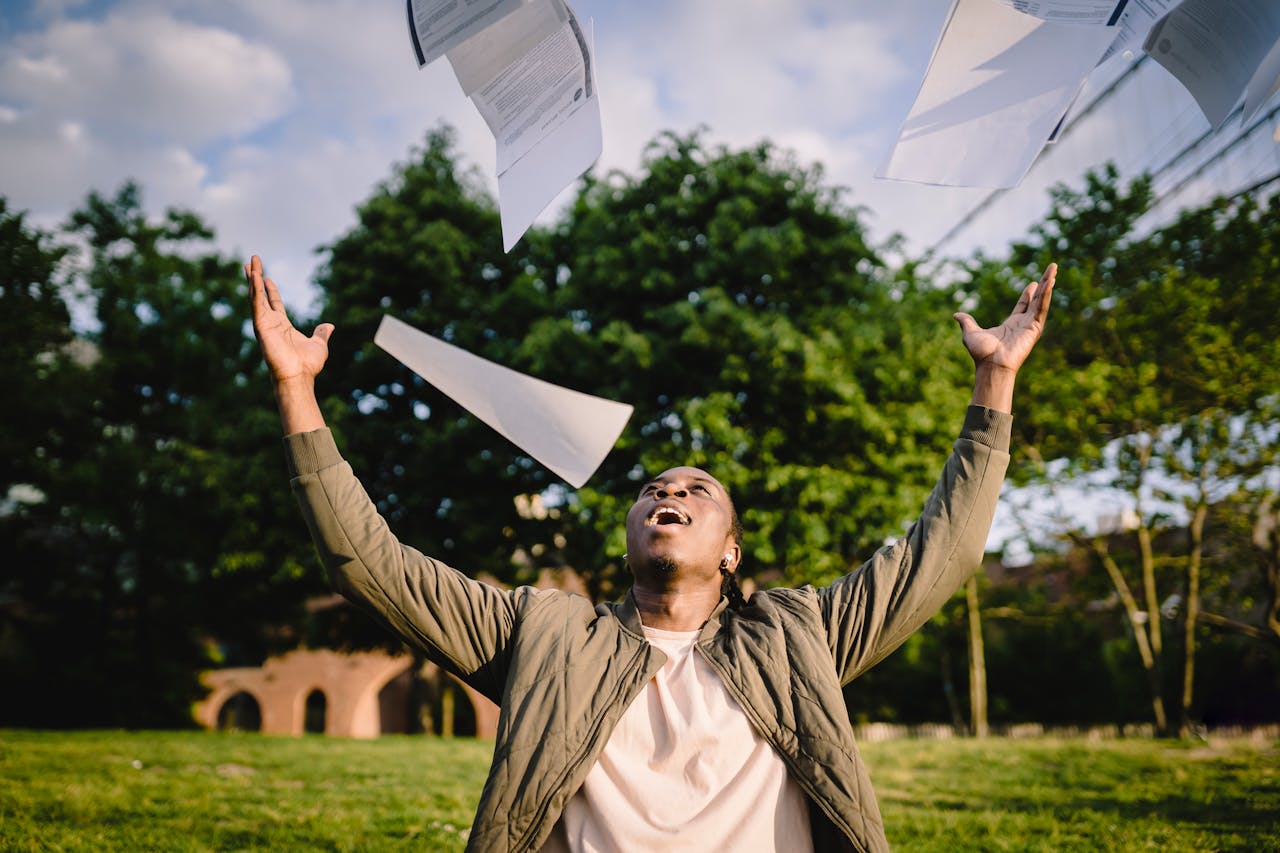 Cheerful young African American male student in casual clothes throwing college papers up in air while having fun in green park after end of exams